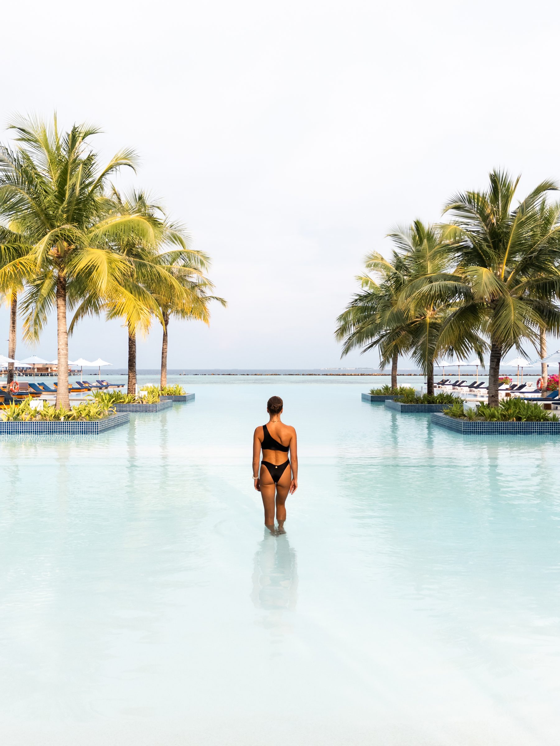 Woman walking into a white infinity pool lined with palm trees, Maldives resort